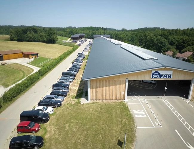Widok z lotu ptaka na budynek z parkingiem zlokalizowanym wzdłuż elewacji. Na parkingu zaparkowane są samochody, a obiekt otaczają pola i zieleń.