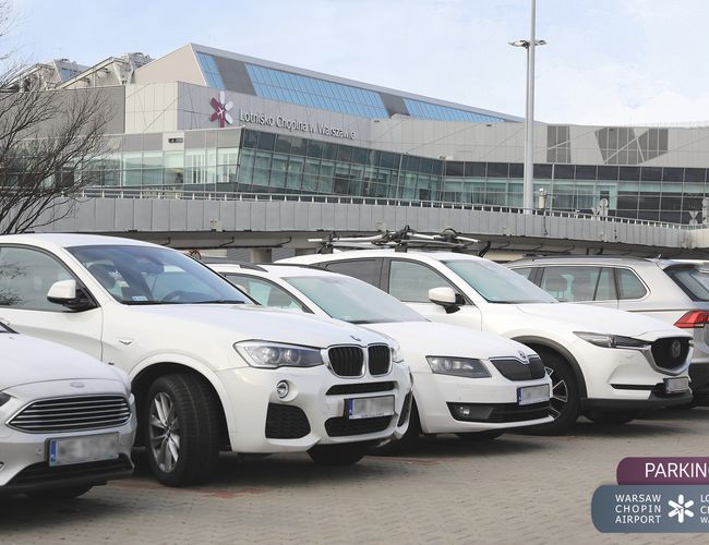 Oficjalny Parking lotniska P4 - parking jest pełen samochodów. Budynek lotniska widoczny w tle. Teren jest otwarty i niezadaszony.