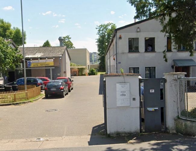 Parking ParkPoint24 Valet - parking z żwirowaną nawierzchnią. Ogrodzony płotem, otoczony drzewami. Obok stoi budynek.
