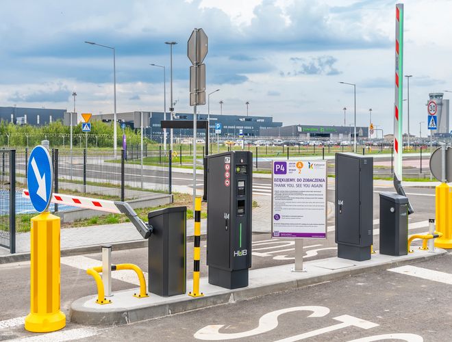 Oficjalny Parking P4 – Wjazd z szlabanem i automatem biletowym. Widoczne oznakowanie poziome i znaki drogowe. Teren jest ogrodzony.