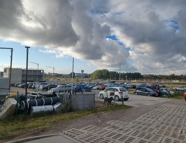 Parking Goliat – plac jest otoczony siatką. Widać zaparkowane samochody. Na horyzoncie stoją dźwigi budowlane.
