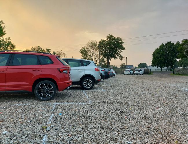 Parking MPark - Coches diversos están estacionados en la superficie de grava. Al fondo hay árboles y una puerta. El aire es tranquilo.