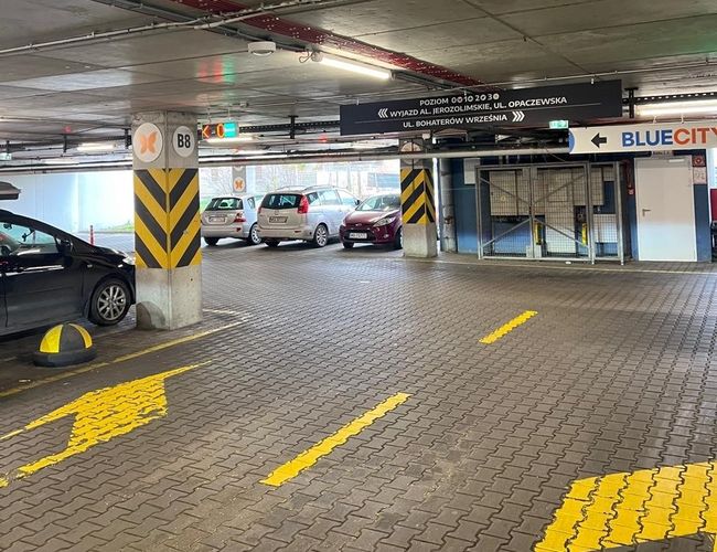 Parking P2 - Okęcie Północ Wielopoziomowy - Widać wnętrze parkingu z żółtymi liniami. Kilka samochodów stoi zaparkowanych w rzędzie.