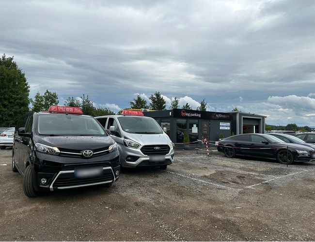Kraków KING parking – Dwa auta zaparkowane przed nowoczesnym budynkiem. Parking jest na żwirowym placu z wyznaczonymi miejscami. Ograniczony taśmą.