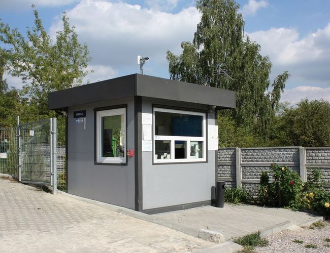 Parking Pienista 32 – mały budynek administracyjny przy ogrodzeniu. Obok rosną drzewa i krzewy. W tle widać betonowy płot.