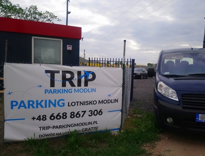 TRIP Parking - Lugar cerrado con caseta y coche. En la reja hay una gran pancarta informativa. Al fondo se pueden ver varios coches aparcados.