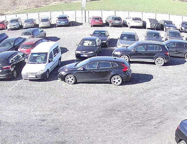 TRIP Parking - Na parkovisku stojí veľa áut. Areál je ohradený a osvetlený. Na pozadí je vidieť zelenú krajinu.