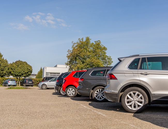 Parking Letim Brno – na zdjęciu kilka zaparkowanych samochodów. Teren jest otoczony drzewami i budynkiem. Pogodny, słoneczny dzień.