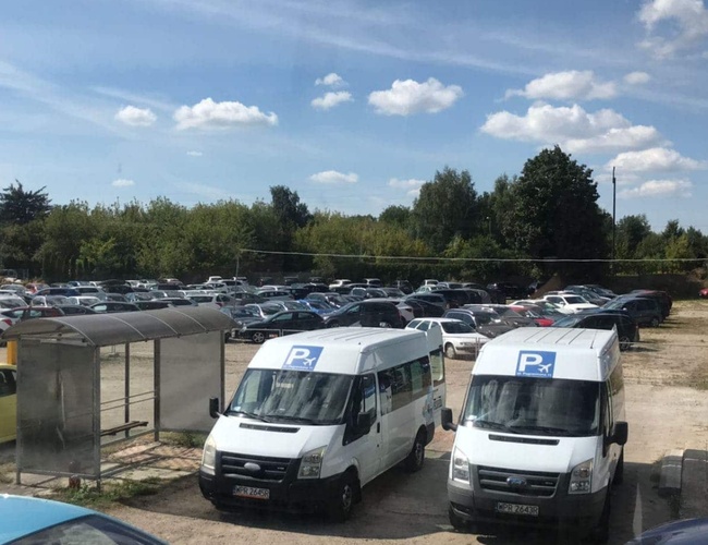 Parkuj Leć - Na parkingu stoją liczne samochody. Obok znajdują się dwa białe busy. W tle jest zieleń i krzewy.