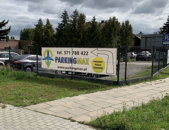 A1 Parking Max – Ogrodzony parking z tablicą informacyjną przy wejściu. Teren z kostki brukowej otoczony jest drzewami.