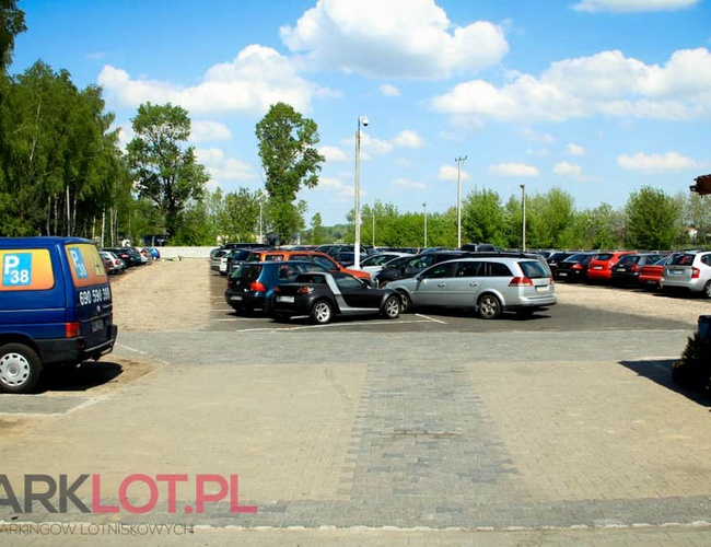 Parkovisko P38 - Pozemok je otvorený a priestranný. Automobily stoja v jednoduchých radoch. V pozadí sú vidieť stromy.