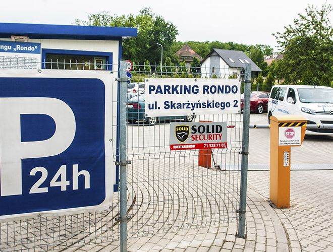 Parking Rondo – ogrodzony teren z bramą wjazdową. Jest tablica informacyjna. W tle stoją samochody.