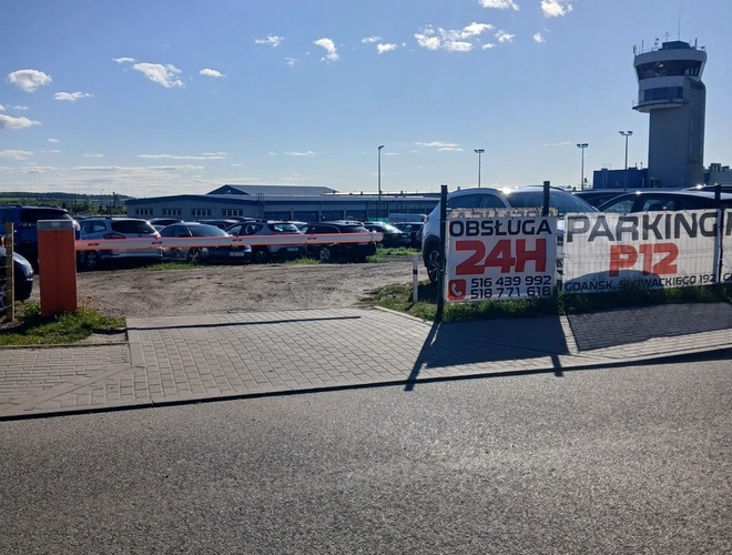 Parking P12 – Na parkingu stoją samochody. Widoczny jest baner z napisem "Obsługa 24H". W tle widać budynek z wieżą.