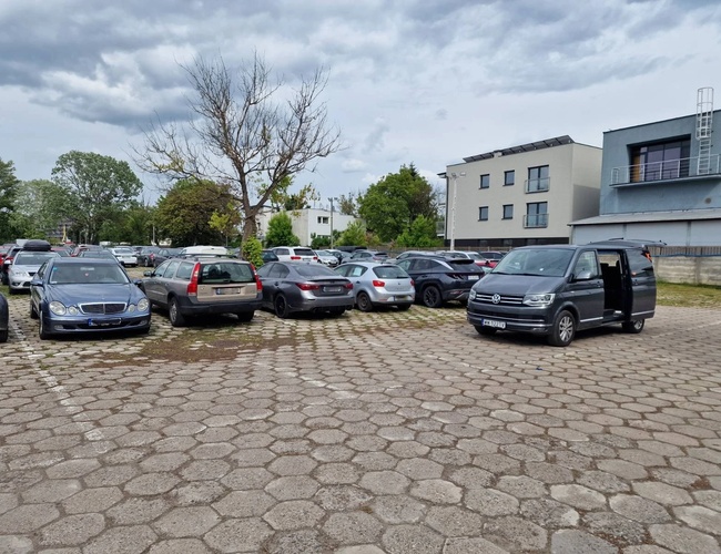 Widok samochodów zaparkowanych na parkingu z samochodem transferowym. Utwardzony kostką teren.