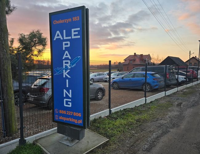 AleParking zadaszony - Parking ogrodzony siatką. W tle widać samochody. Obok stoi duży niebieski szyld.