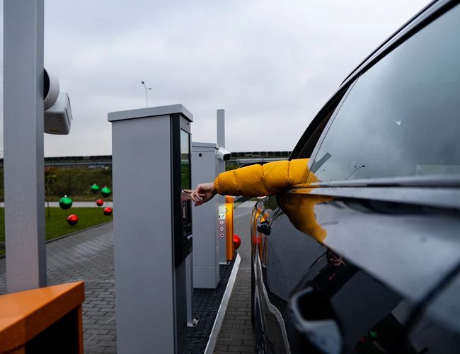 Easy Parking Standard - Osoba w żółtej kurtce opuszcza szybę samochodu. Sięga do parkometru stojącego obok pojazdu. Teren jest ogrodzony panelami.