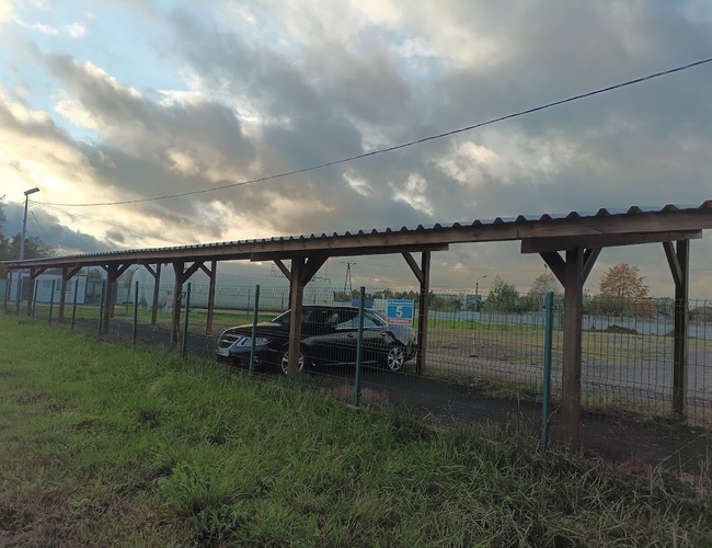 Parking na 5 – zadaszone miejsca postojowe z metalowym ogrodzeniem. Widać kilka aut zaparkowanych pod wiatą. Teren trawiasty wokół jest zadbany.