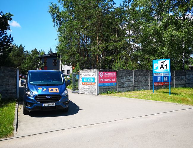 Parking A1 - Wjazd ogrodzony, tablice informacyjne. Samochód zaparkowany przed bramą. Drzewa w tle.
