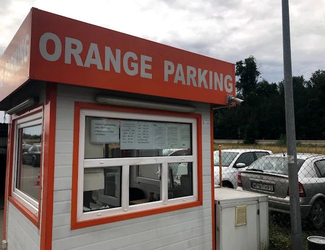 Aparcamiento Naranja Modlin - Edificio visible con letrero y ventanas. En el fondo hay coches estacionados. El área está cercada con una malla.