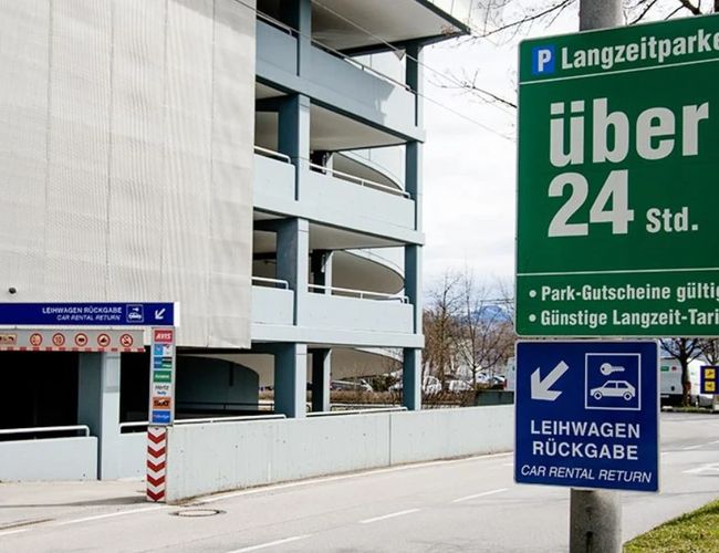 Wjazd do wielopoziomowego parkingu z wysokim znakiem informacyjnym umieszczonym przed budynkiem. Obok widoczna jest droga prowadząca do parkingu.