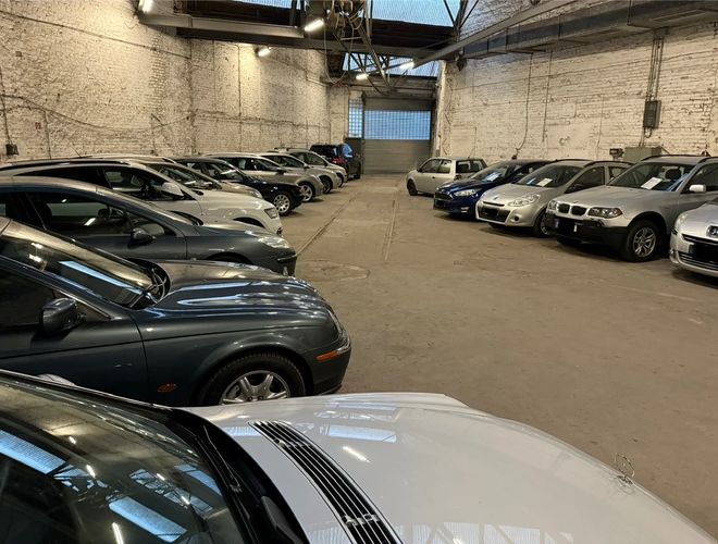 Parking Park Elite Valet - Wnętrze hali z zaparkowanymi autami. Samochody stoją w dwóch rzędach. Widać duże, metalowe drzwi z tyłu.