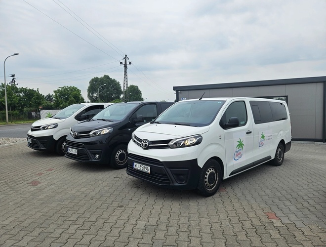 Holiday Parking - Holiday Parking: tres vehículos aparcados uno al lado del otro. Los dos blancos tienen el logotipo con una palmera. El aparcamiento está pavimentado y vallado.