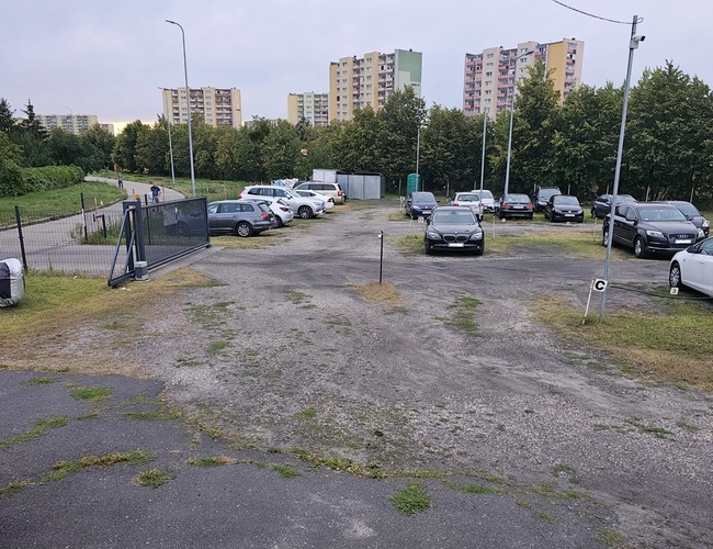 Odlotowy Parking BZG – Parking jest ogrodzony metalowym płotem. Na placu stoją zaparkowane samochody. W tle widać bloki mieszkalne.