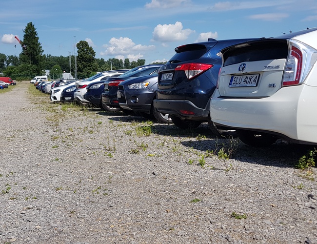 DOBRÉ Parkovisko - Štrkový povrch s zaparkovanými autami po oboch stranách. V pozadí sú viditeľné stromy a obloha s oblakmi.