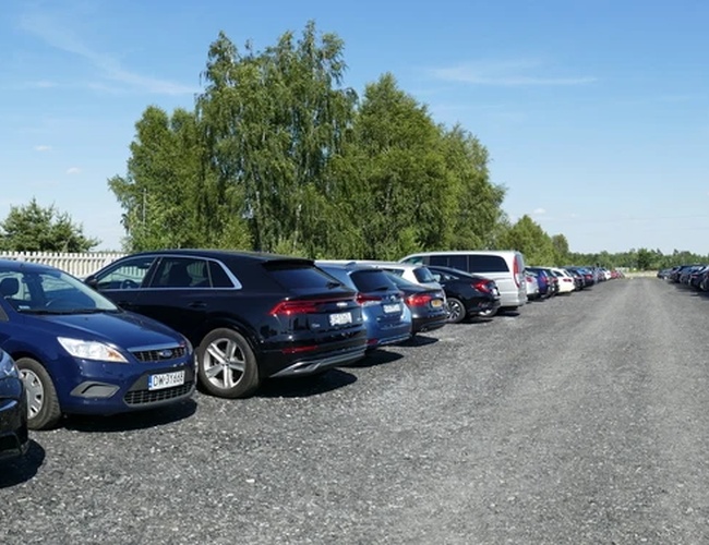 Diza Parking - En el área de grava están aparcados coches. Al fondo se ven árboles. El clima es soleado.