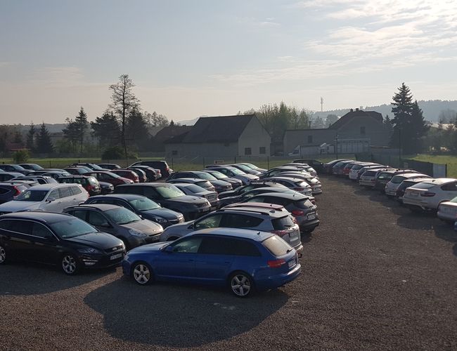 BaliceLot24 – Widoczny jest duży, nieutwardzony parking z wieloma samochodami. Teren otoczony płotem i sąsiedztwo zieleni dodają otwartości.