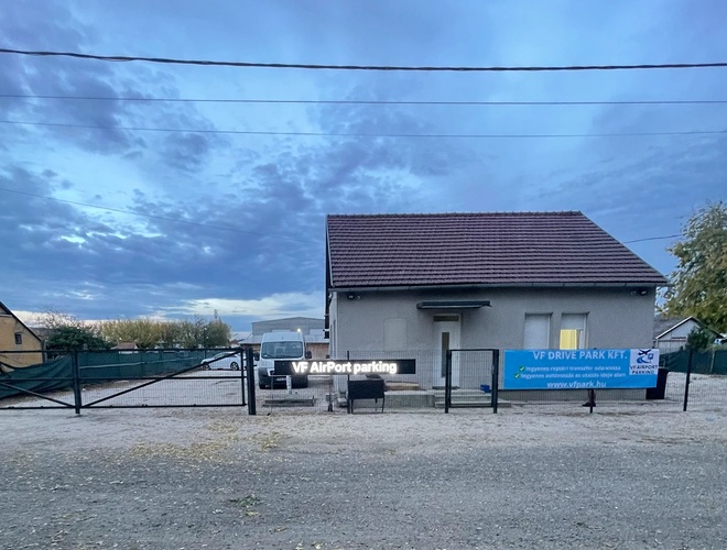 Mały, szary budynek z czerwonym dachem na terenie żwirowego parkingu oznaczonego jako ‘VF AirPort parking’. Na ogrodzeniu widoczny jest baner z napisem ‘VF Drive Park Kft.’ oraz adres strony internetowej. W tle widać zaparkowany biały samochód dostawczy i