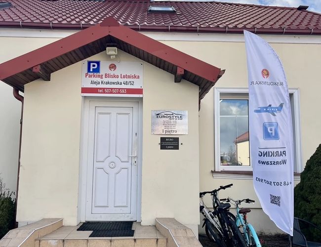 Parking Blisko Lotniska - Wejście do budynku z białymi drzwiami i schodami. Obok znajduje się stojak z rowerami i biały baner.