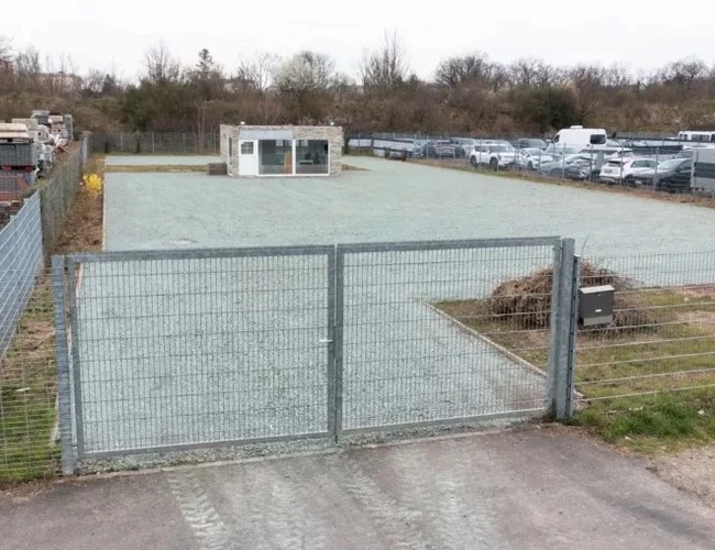 Estacionamiento de transporte - El área está cercada por una valla de metal. En el medio se puede ver un pequeño edificio blanco. A los lados hay coches estacionados.