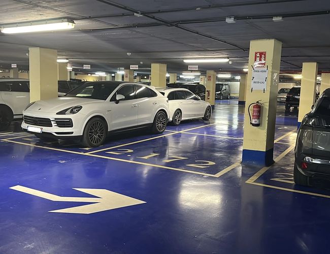 Parcare Mașină și Zbor Garage Valet - garaj închis cu câteva mașini. Pardoseala este albastră cu marcaje de locuri. Luminile de tavan iluminează interiorul.