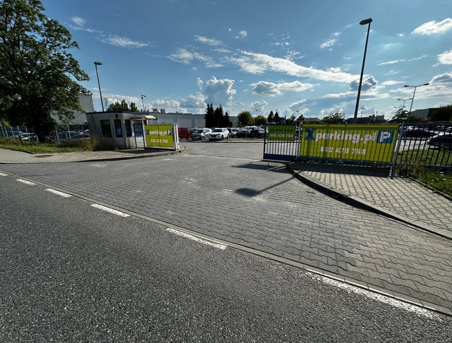 Widok na bramę wjazdową parkingu. W tle widok samochodów zaparkowanych na parkingu, którego teren jest wyłożony kostką brukową.