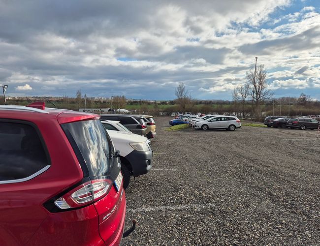 HostParking - Parking żwirowy z zaparkowanymi samochodami. Teren nie jest ogrodzony. W tle widoczne drzewa i chmury.