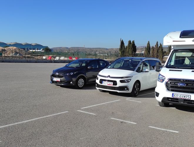 Parkplatz Odkryj-Auto - Drei Fahrzeuge auf offenem Gelände geparkt. Die Autos stehen nahe beieinander. Im Hintergrund sind Berge und ein Zaun sichtbar.