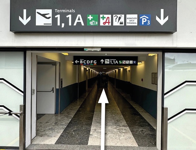 Parking Parktiger Transfer – Entrada a un largo pasillo. Arriba señales direccionales. Suelo con patrón de franjas.