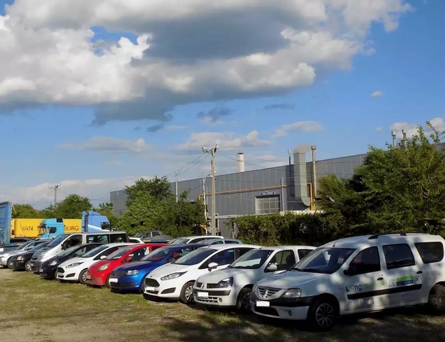 Rząd kolorowych samochodów zaparkowanych przy trawniku obok budynku przemysłowego w słoneczny dzień.