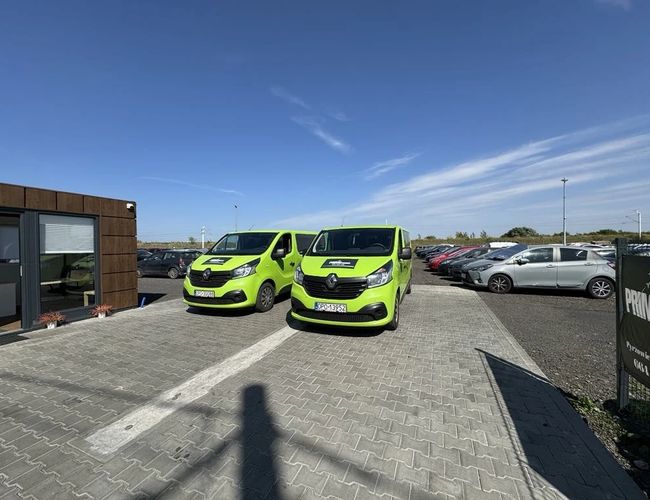 Parking Prime - Zwei grüne Vans stehen auf der gepflasterten Straße. Auf der rechten Seite sind Autos geparkt.