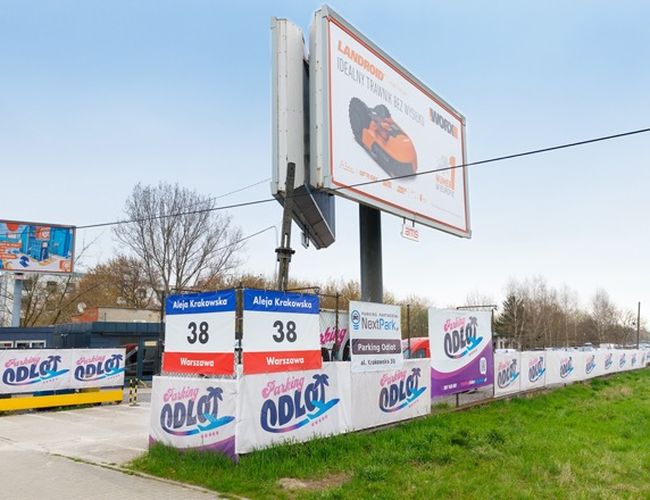 Parking Odlot – Widać wejście z ogrodzeniem i reklamą. Tablice informacyjne wskazują lokalizację. Całość otoczona jest zielenią.