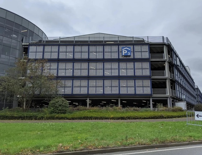 Zewnętrzny widok na nowoczesny parking wielopoziomowy z przeszkloną fasadą, położony w pobliżu terminala lotniska i głównych dróg dojazdowych.