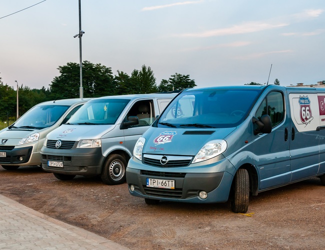 AAP-66 – Na parkingu stoją trzy dostawcze samochody. Każdy pojazd ma widoczne logo. W tle widać zielone drzewa.