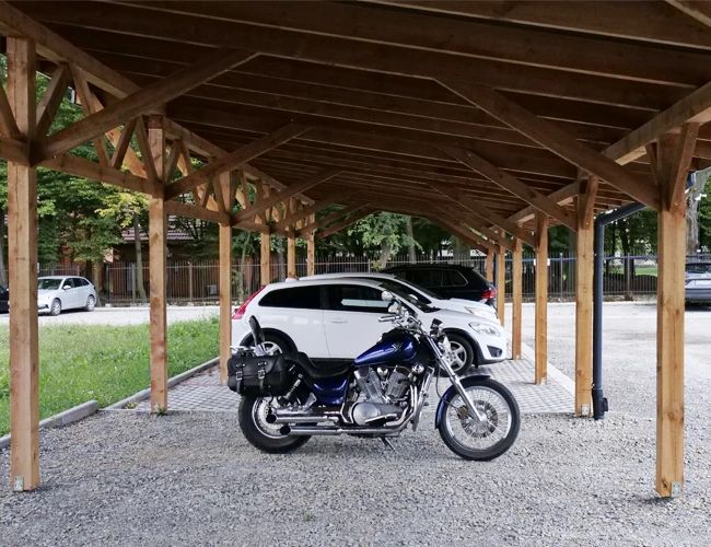 Parking Zielony Kompleks Dwór Ostoya - Zadaszony parking z motocyklami i samochodami. Drewniana konstrukcja zapewnia ochronę. W tle widać ogrodzenie i drzewa.