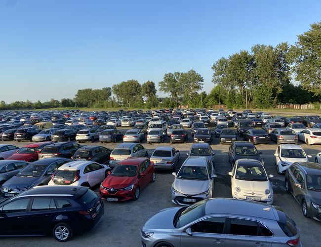 Odkryj-Parking – Na parkingu stoi wiele samochodów. Teren jest otwarty i otoczony drzewami. Pogoda jest słoneczna.