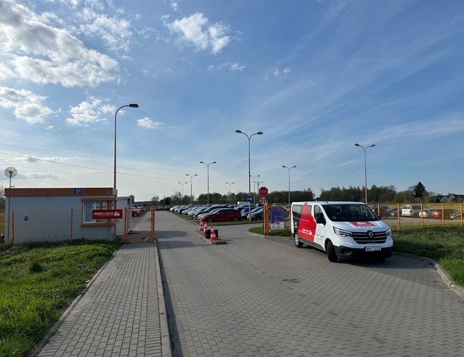 Objavte - Parkovisko GDN – Parkovisko je oplotené a osvetlené. Stojí pri ňom biely dodávkový voz. Nachádza sa blízko vjazdu na pozemok.