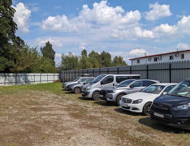 Parking P60 – kilka samochodów stoi na żwirowej nawierzchni. Teren jest ogrodzony metalowym płotem. W tle rosną drzewa i widać budynek.