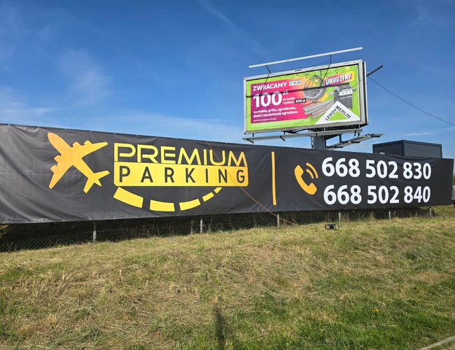 Prémiové parkovisko - Baner s logom a telefónnymi číslami. Vedľa je billboard s reklamou stavebnín. Pozemok je oplotený sieťou.