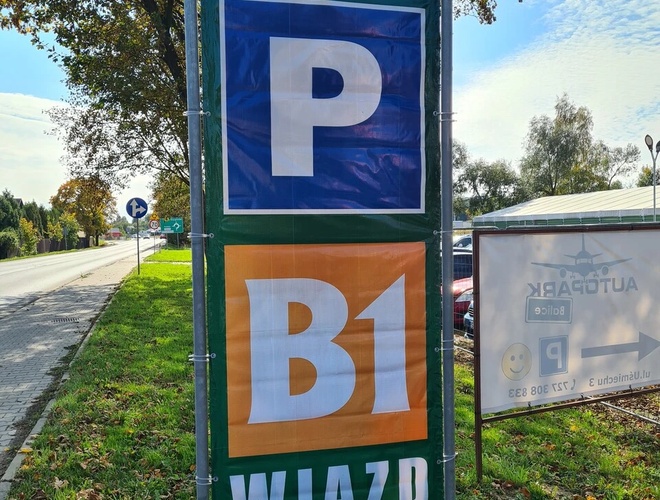 B1 Parking – Dwa dostawcze samochody stoją przed ogrodzeniem. Obok są fabryczne budynki. Na horyzoncie widać turbinę wiatrową.