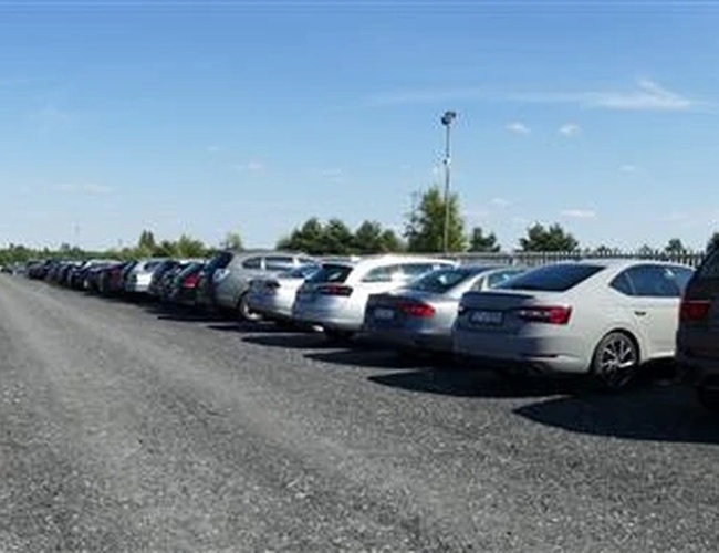 Diza Parking – En el aparcamiento hay coches estacionados en dos filas. El terreno es de grava y está rodeado de vegetación. Se puede ver iluminación.